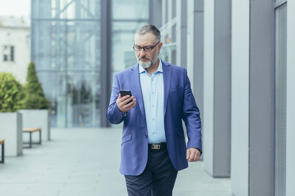 Serious gray-haired senior handsome man businessman in suit and glasses walking near office center on street, holding mobile phone, typing message, checking mail. - 写真・画像
