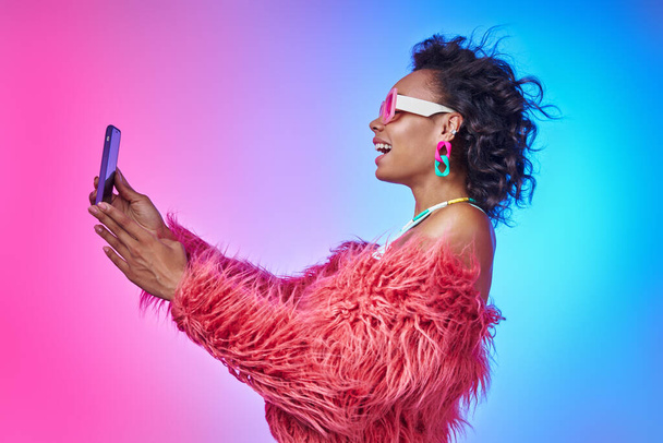Fashionable African woman in fluffy jacket holding smart phone and smiling against colorful background - Zdjęcie, obraz