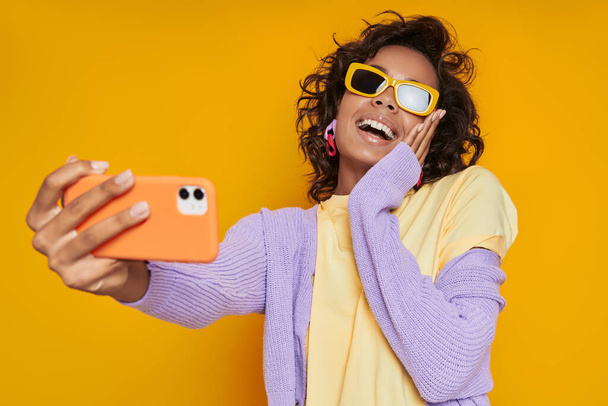 Beautiful African woman in funky eyeglasses making selfie and smiling against yellow background - Fotografie, Obrázek