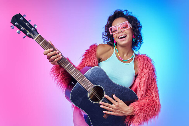 Attractive African woman in fluffy jacket playing guitar and smiling against colorful background - Фото, изображение