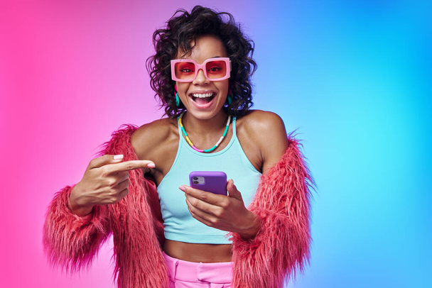 Excited African woman in trendy clothes pointing her smart phone and smiling against colorful background - Fotografie, Obrázek
