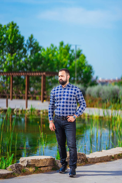 a bearded man stands in a park next to a pond. Turkmen guy is resting in the park. photo session of an office worker in the park - Fotoğraf, Görsel