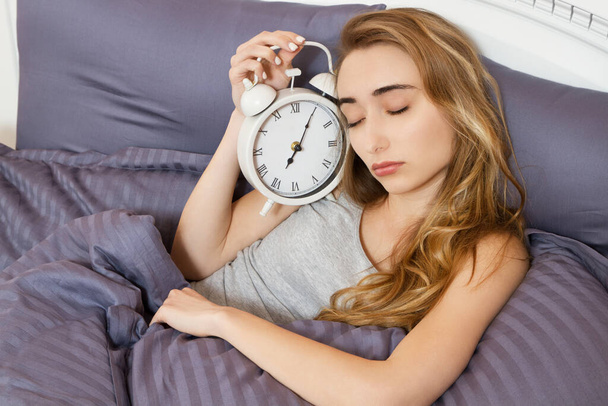 Mladá krásná ospalá dívka se zavřenýma očima drží hodiny a leží ve své posteli v ložnici v dopoledních hodinách - Fotografie, Obrázek