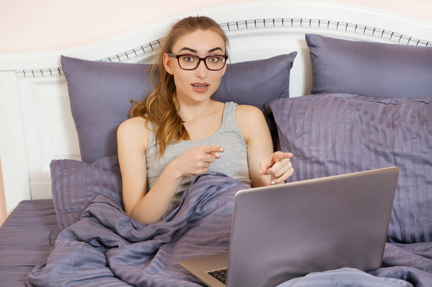 Pretty teen woman using her laptop in bed. Beautiful girl in glasses working on laptop in bedroom - Fotografie, Obrázek