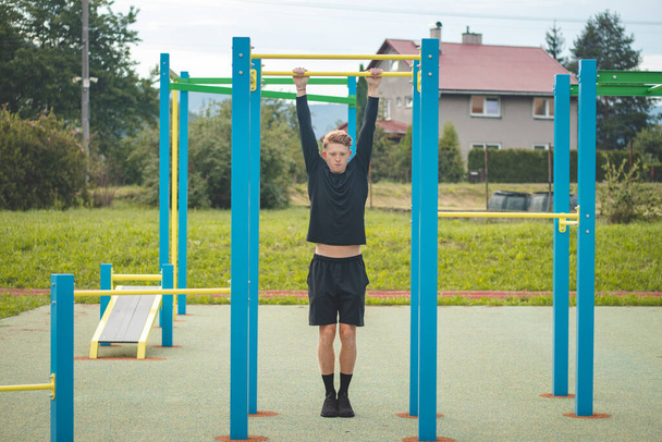 El chico rubio orientado a las metas construye la fuerza de su propio cuerpo en el campo de entrenamiento durante el verano. Tríceps de entrenamiento, bíceps, hombros y abdomen. Crosfit ejercicio pull-ups. - Foto, Imagen