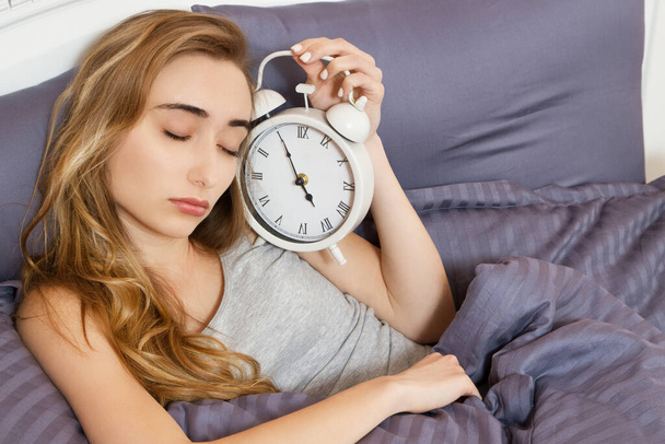 Молодая красивая сонная девушка с закрытыми глазами держит часы и лежит в постели в спальне утром - Фото, изображение