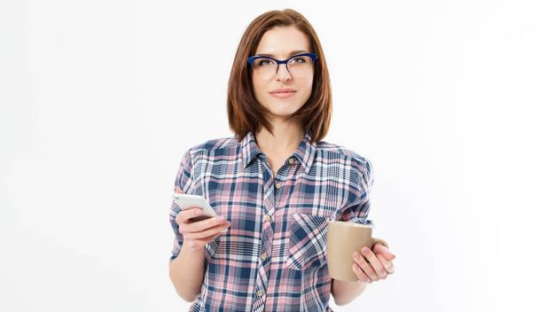 Pretty young woman holding smart phone, using device, wearing stylish glasses, smiling, holding a mug, isolated on white background, denim shirt - Photo, Image