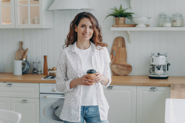 Smiling girl holding paper cup of coffee standing in modern cozy kitchen, looking at camera. Happy female housewife homeowner holds takeaway delivery hot drink to go at home. Domestic life - 写真・画像