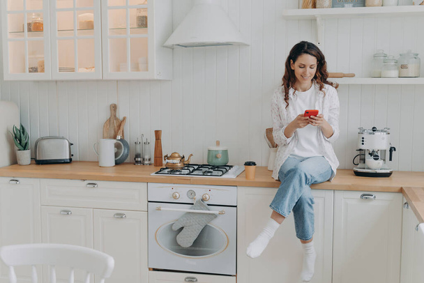 Szczęśliwa kobieta gospodyni siedzi w nowoczesnej, czystej i przytulnej kuchni w domu ze smartfonem. Uśmiechnięty klient e-commerce kobiet zakupy online, cieszyć się odpoczynku po pracach domowych, zadowolony z usług sprzątania domu. - Zdjęcie, obraz