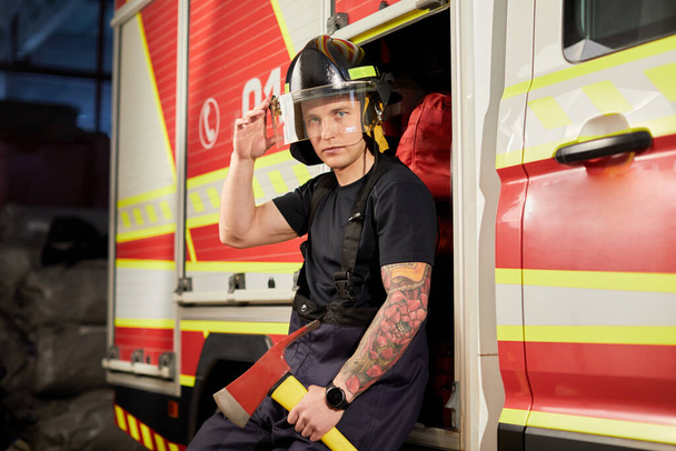 消防士の消防車に対する斧付きヘルメットの写真.  - 写真・画像