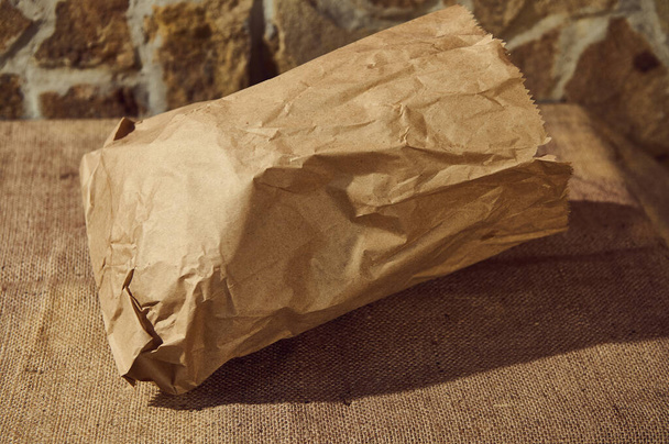 Close-up of an eco-friendly recyclable craft paper bag with healthy wholegrain wheat bread inside, on a table with a sackcloth tablecloth. Copy advertising space. Selective focus - Foto, Imagem