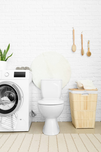 Современный унитаз, корзина для белья и стиральная машина возле стены белого кирпича в ванной комнате - Фото, изображение