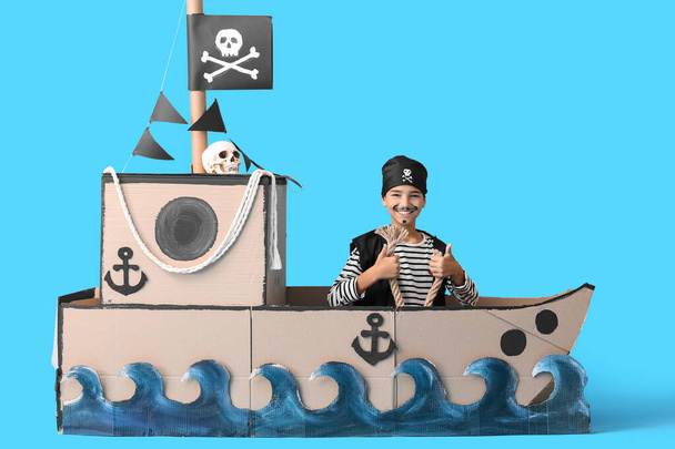 Little boy dressed as pirate with rope in cardboard ship on blue background - Photo, Image