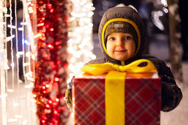 Criança feliz em um chapéu vermelho de Papai Noel com presente de Natal embrulhado perto de lâmpadas brilhantes na noite de inverno. O conceito de Natal e Ano Novo. - Foto, Imagem