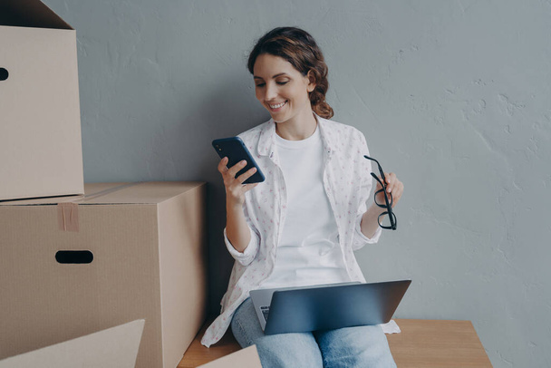 Happy businesswoman using app on mobile phone, ordering parcel delivery. Female office manager preparing online store orders in carton boxes for sending. E-commerce, small business concept. - Photo, Image