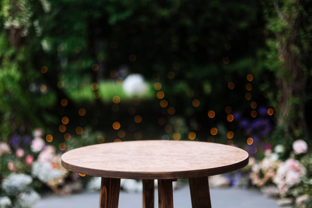 Round wooden table on a green nature with light bulbs background. Outdoor table at desk. Grass, greens and party decoration bushes in the background - Photo, image