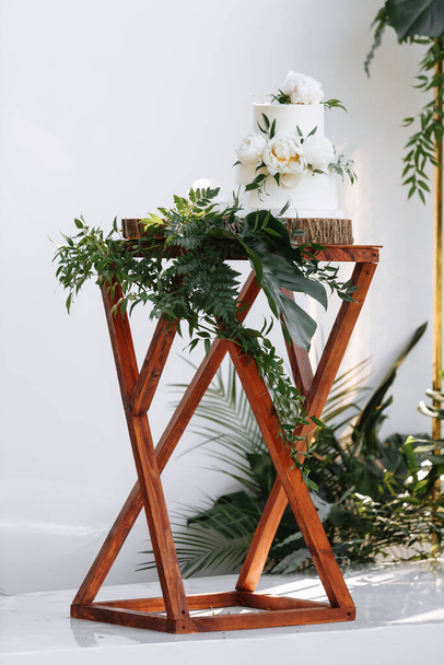 White two-tiered wedding cake decorated by white peonies flowers and greenery with stand on wooden table decoration monstera leaf. Wedding. Reception. - Φωτογραφία, εικόνα