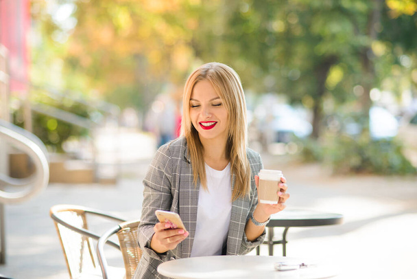 πορτρέτο μιας ξανθιάς γυναίκας με όμορφο χαμόγελο, που κάθεται σε ένα τραπέζι σε ένα καφέ του δρόμου, με ένα τηλέφωνο και καφέ στα χέρια της - Φωτογραφία, εικόνα
