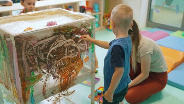 Cling film painting. Toddler painting with a sponge, brushes and paints on a cling film wrapped all the way round the wooden shelf unit. A teacher helping them. Creative activity for kids sensory - Video