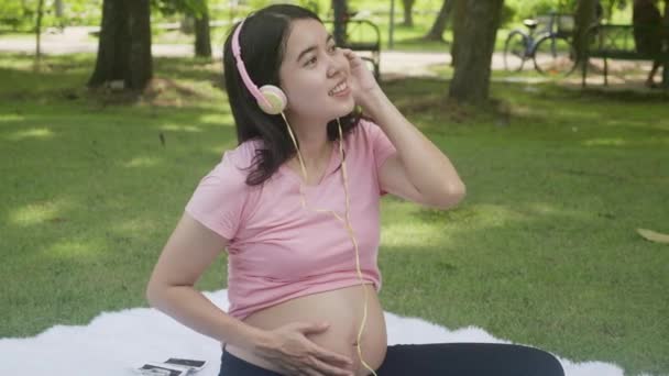 Happiness young asian woman pregnant sitting and listening music with for relaxation in the park, mother leisure with positive in the garden, activity for pregnancy and lifestyles concept. - Felvétel, videó