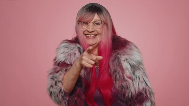Эй ты. Женщина 70 лет в пальто радостно улыбается и указывает на камеру, выбирая счастливого победителя лотереи, указывая на удивительный вас. Пожилая бабушка изолирована в одиночестве на фоне розовой стены студии - Кадры, видео