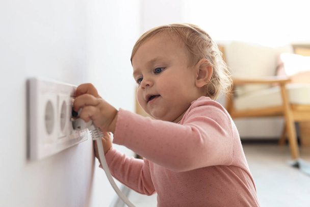Cute baby playing with electrical socket and plug at home. Dangerous situation - Foto, afbeelding