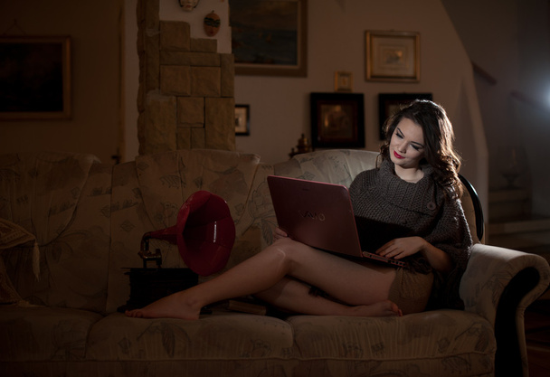 Beautiful young woman sitting on sofa working on laptop having a red gramophone near her, in boudoir scenery. Attractive brunette girl with long hair and long legs laying down on couch with a laptop - Фото, изображение