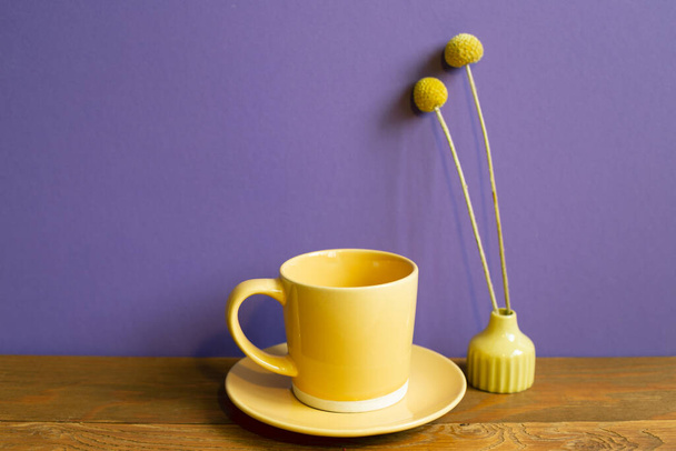 Žlutý šálek kávy a suchý květ na dřevěném stole. fialové pozadí zdi. retro styl - Fotografie, Obrázek