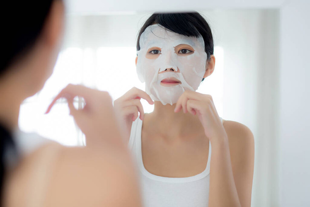 シートフェイスマスクと家で鏡を見て美しい若いアジアの女性,しわのためのスキンケア顔と化粧品や化粧品を適用する美少女,治療と保湿剤,健康と健康. - 写真・画像