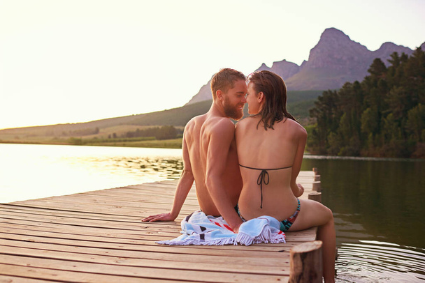 Idling away the day. Rearview shot of an affectionate young couple in swimsuits sitting on a dock at sunset - Photo, Image