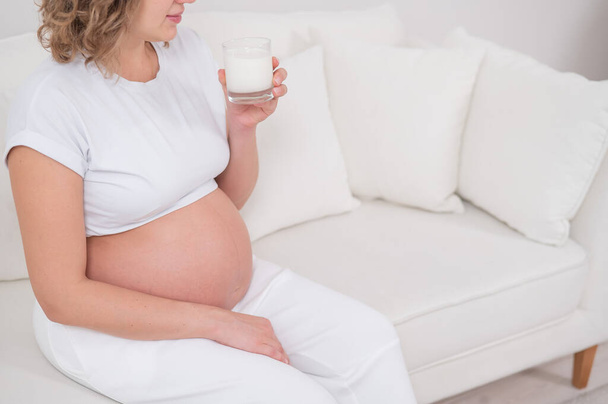 A pregnant woman sits on a white sofa and drinks milk - Fotoğraf, Görsel