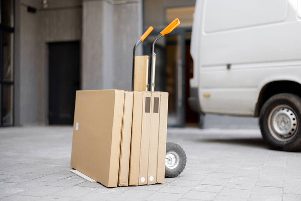 Folding furniture in cardboard boxes on a pushcart delivered by vehicle near building entrance. Concept of home delivery - Foto, Bild