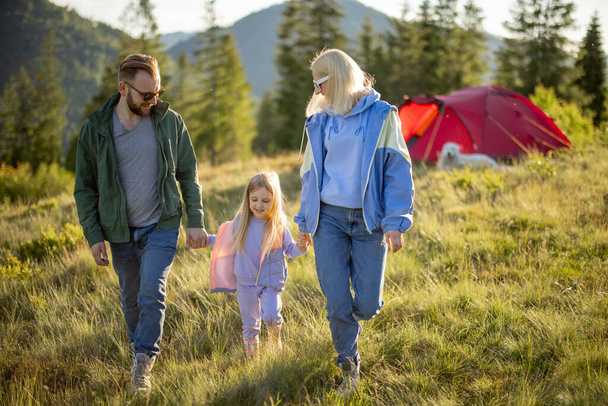 Jonge Kaukasische familie met klein meisje lopen samen op groene weide tijdens het reizen met tent in de bergen. Gelukkige familie zomervakantie op camping - Foto, afbeelding