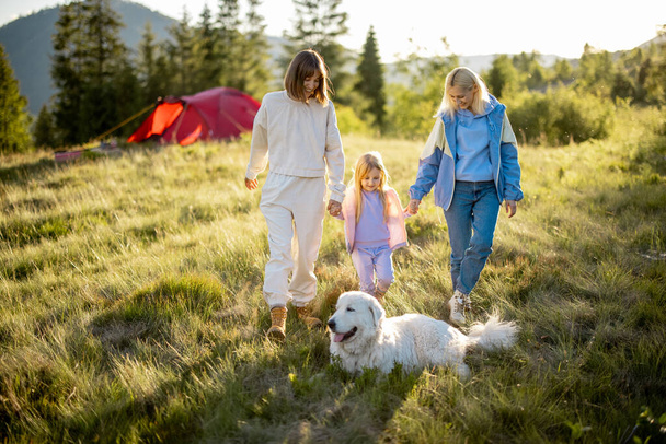 Δύο γυναίκες περπατούν με το μικρό κορίτσι και το άσπρο σκυλί, περνώντας ευτυχισμένα το καλοκαίρι στη φύση. Ομοφυλόφιλος οικογένεια με ένα παιδί ταξιδεύει στα βουνά - Φωτογραφία, εικόνα