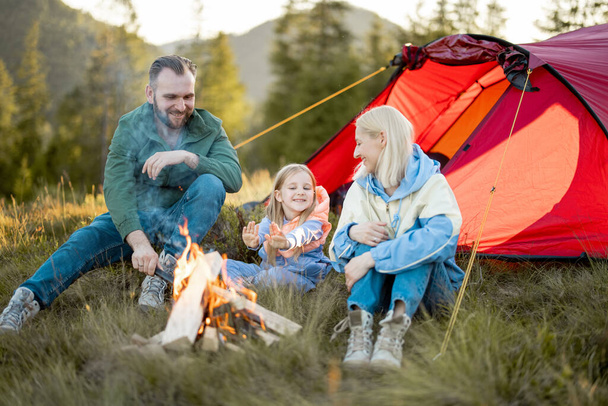 Young adult couple sit with a little girl and have lovely conversation by the campfire, traveling with tent on nature. Family spend summer time hiking in the mountains - Photo, Image