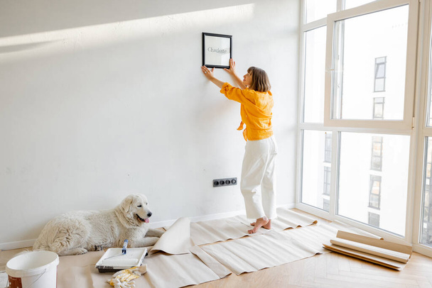 Young woman hanging picture frame in room, decorating her newly renovated apartment, stands with her dog in white room - Foto, afbeelding