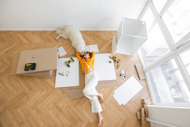 Young woman lies on floor with her cute dog, resting while making repairing at new apartment, top view. Young woman assembling furniture by herself. DIY concept - Zdjęcie, obraz