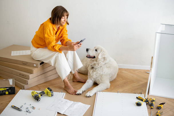 Young woman sits with her cute dog, using phone during repairing at new apartment. Assembling furniture by herself. DIY concept - Foto, immagini
