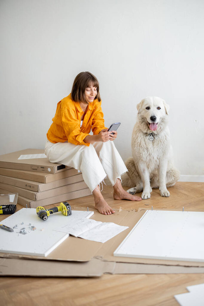 Young woman sits with her cute dog, using phone during repairing at new apartment. Assembling furniture by herself. DIY concept - Foto, Bild