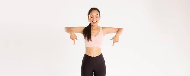 Sport, wellbeing and active lifestyle concept. Excited and happy asian fitness girl in sportswear, pointing fingers down and looking amazed with great discounts, read banner, white background. - Photo, Image