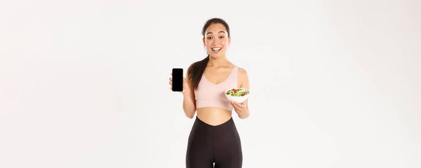 Sport, wellbeing and active lifestyle concept. Smiling slim and cute asian fitness girl, gym coach showing salad and smartphone screen, recommend download diet tracker or calories reminder. - Photo, Image