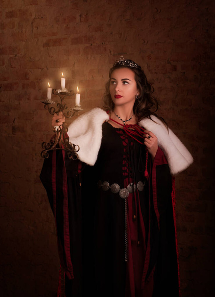 Fantastic medieval woman with a candlestick in her hands. Royal velvet vintage red dress with fur and gold, princess, queen, brunette with long hair. Castle, dusk, night, luxury - Fotó, kép