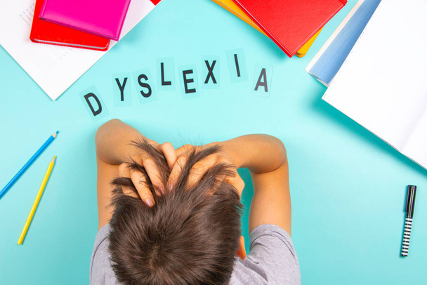 Sad tired frustrated teenage boy sitting at table with many books and school supplies. Word Dyslexia on light blue background. Dyslexia, learning disability, reading difficulties, education concept. - Photo, Image