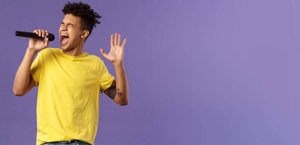 Portrait of passionate carefree young hispanic singer with dreads and tattoos, reaching highest note in song, raising hand up singing loud at microphone with closed eyes, purple background. - 写真・画像