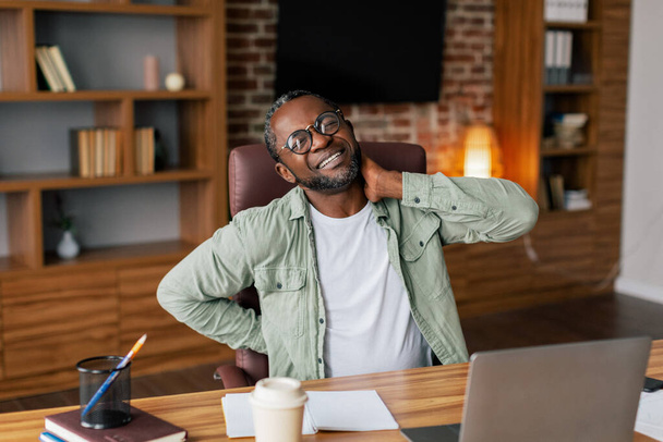 Trieste volwassen Afro-Amerikaanse man manager in bril, casual lijdt aan pijn in rug en nek van stress en overwerken op laptop in het thuiskantoor interieur. Gezondheidsproblemen, gezichtsuitdrukking en pijn - Foto, afbeelding