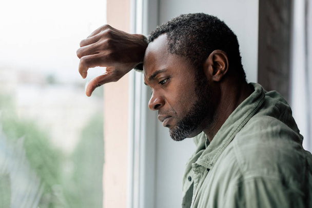 Unglücklich frustrierter erwachsener afrikanisch-amerikanischer Kerl in lässiger Weise, der an Depressionen und schlechten Nachrichten in der Nähe von Fenstern im häuslichen Innenraum leidet. Gesundheitliche Probleme, Stress durch Selbstisolation während der Covid-19-Quarantäne - Foto, Bild