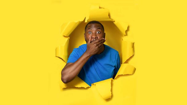 Unbelievable news. Shocked black man closing mouth with hand in surprise, looking through hole in torn yellow paper. Omg, great offer, cool promotion, amazing news - Photo, Image