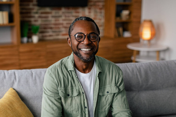Headshot του χαρούμενου ενήλικα Αφροαμερικανός άνδρας με γυαλιά και casual χαλάρωση στον καναπέ και κοιτάζοντας κάμερα στο εσωτερικό του σαλονιού, κλείστε. Επιχειρήσεις και εργασία στο σπίτι κατά τη διάρκεια της καραντίνας covid-19 - Φωτογραφία, εικόνα