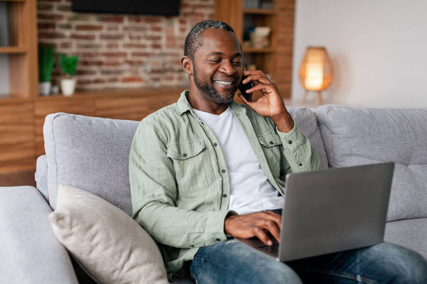 Smiling elfoglalt középkorú afro-amerikai üzletember alkalmi hívás telefonon és gépelés laptop a nappali belsejében. Szabadúszó, munka és üzleti otthon, új normál és társadalmi távolság - Fotó, kép