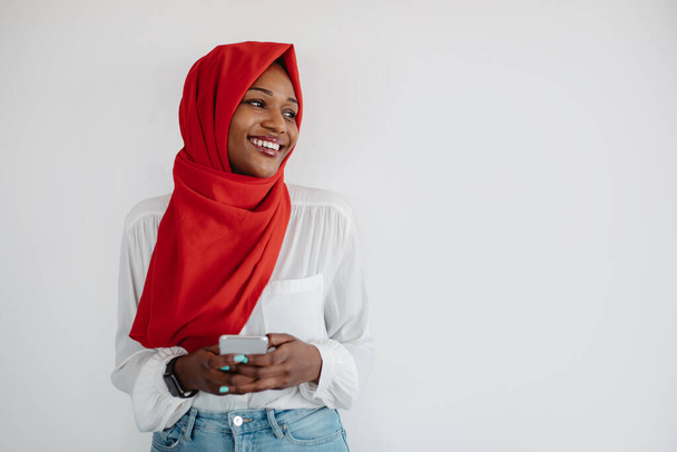 Szczęśliwy muzułmanin czarna kobieta w chustce za pomocą smartfona i patrząc na wolną przestrzeń nad jasnym tle ściany. Afrykańska amerykanka demonstruje przestrzeń na tekst lub projekt - Zdjęcie, obraz